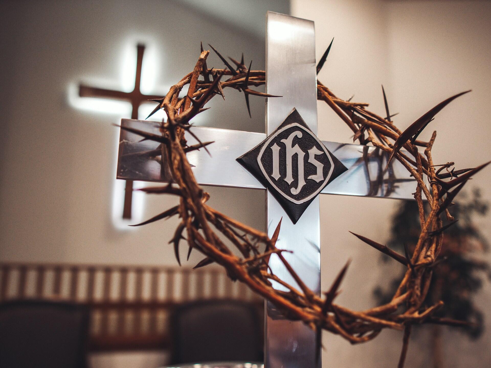 A close-up of a silver cross encircled by a thorn crown, featuring the letters "IHS" on a dark diamond background.